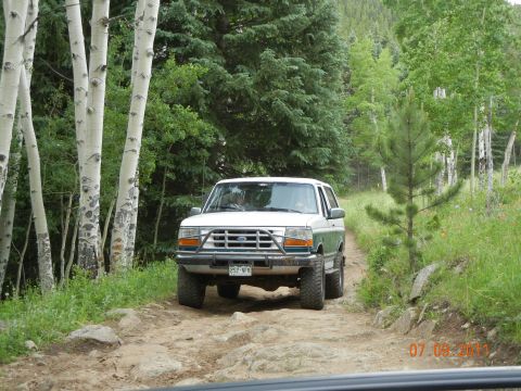 1996 Ford Bronco - A white 1996 Ford Bronco navigates a rocky dirt road surrounded by trees.