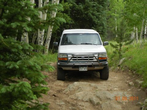 1996 Ford Bronco - White 1996 Ford Bronco navigating a rough, off-road path through trees.