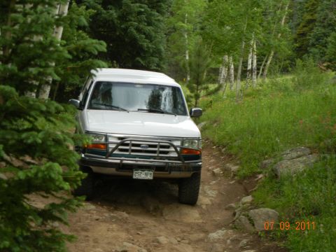 1996 Ford Bronco - A white SUV navigates a rough, wooded trail on a summer day.