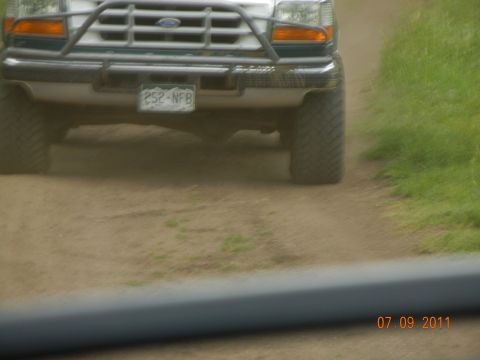 1996 Ford Bronco - A white Ford truck with a brush guard drives on a dirt road, with grass on the right.