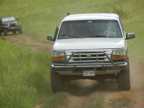 1996 Ford Bronco - White 1996 Ford Bronco drives on a dirt path, with another vehicle visible behind it.
