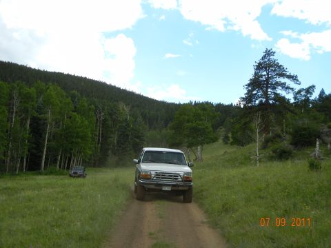 1996 Ford Bronco - Two SUVs drive on a dirt road through a grassy, wooded area under a cloudy sky.