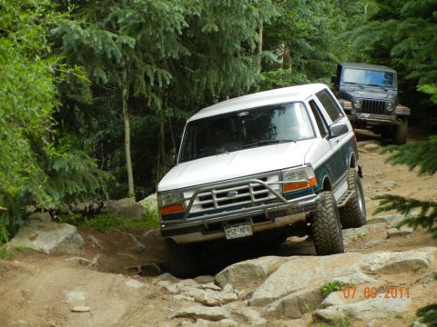 1996 Ford Bronco - Two off-road vehicles navigate a challenging rocky path through a forest.