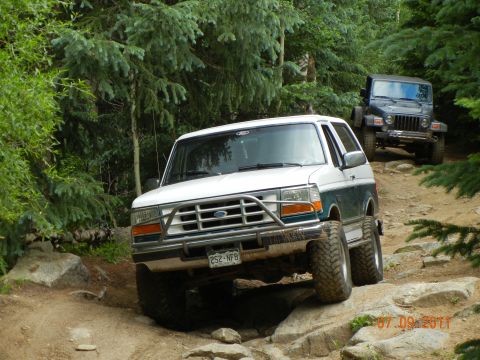 1996 Ford Bronco - Two off-road vehicles navigate a challenging, rocky path surrounded by dense forest.