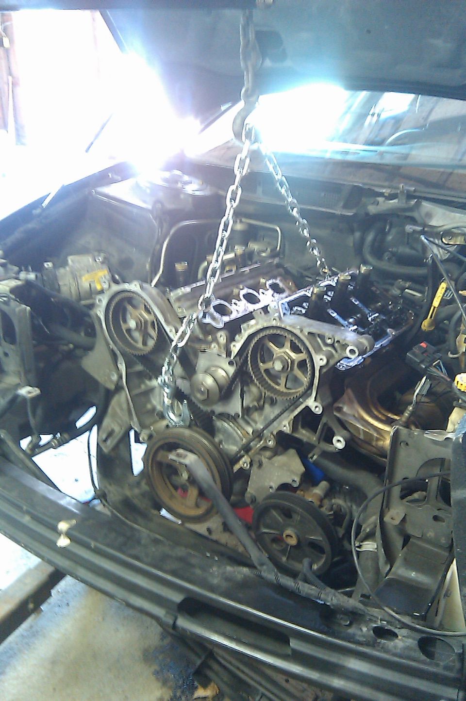 Engine with chains attached, being lifted out of a car. Visible parts include timing belt, pulleys, and engine block