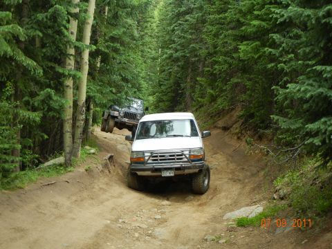 1996 Ford Bronco - Two off-road vehicles navigate a steep, wooded trail on a sunny day.
