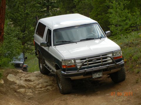 1996 Ford Bronco - A white and dark green Ford Bronco navigates a challenging off-road trail with another vehicle visible behind it.