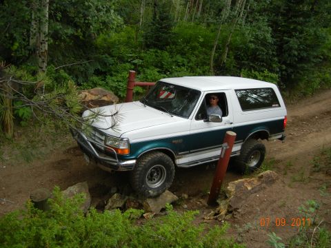 1996 Ford Bronco - A 1996 Ford Bronco is shown off-roading on a dirt path surrounded by dense forest.