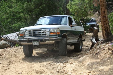 1996 Ford Bronco - Two off-road vehicles navigate a wooded trail with rocky terrain.