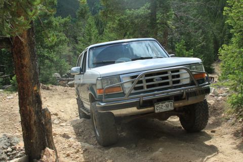 1996 Ford Bronco - 1996 Ford Bronco navigates a rough, dirt path through a forest with trees and rocks.