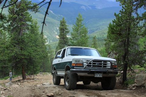 1996 Ford Bronco - This 1996 Ford Bronco is navigating a rocky mountain trail surrounded by pine trees and scenic vistas.