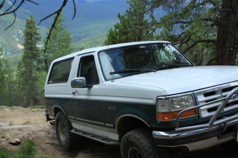 1996 Ford Bronco - This two-tone Ford Bronco is parked on a rocky trail, surrounded by pine trees and a mountainous landscape.