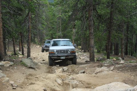 1996 Ford Bronco - Two off-road vehicles navigate a challenging dirt path through a dense forest.