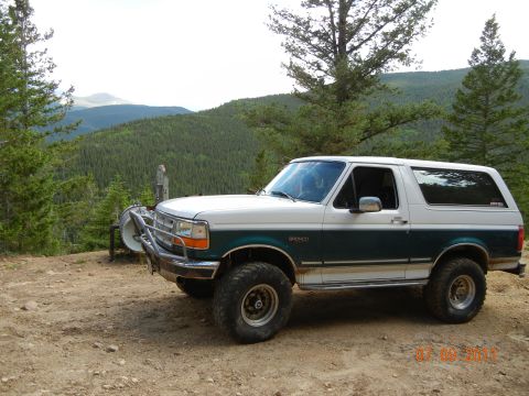 1996 Ford Bronco - A two-tone Ford Bronco sits on a dirt road overlooking a forested mountain range.