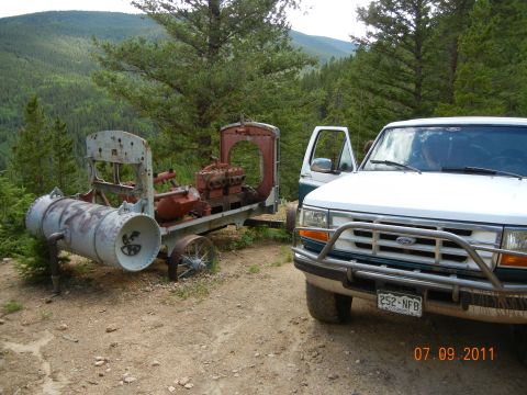 1996 Ford Bronco - A white Ford F-Series truck is parked on a dirt road next to an old piece of industrial equipment in a mountainous area.