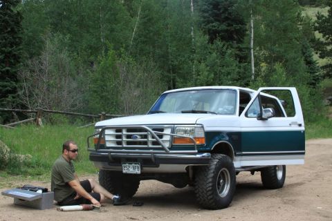1996 Ford Bronco - A man sits next to a lifted 1992 Ford Bronco with its driver's door open, parked on a dirt road with trees in the background.