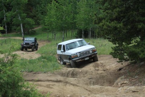 1996 Ford Bronco - A white and gray Ford Bronco navigates an off-road trail, with a black Jeep visible behind it.