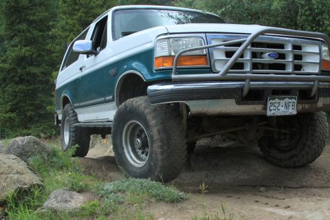 1996 Ford Bronco - This lifted Ford Bronco is navigating a rocky, off-road path with trees surrounding it.