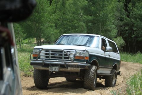 1996 Ford Bronco - A white and green Ford Bronco drives on a dirt road surrounded by trees.