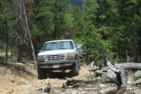 1996 Ford Bronco - A white 1996 Ford Bronco navigates a rocky, wooded trail, with a second vehicle following closely behind.