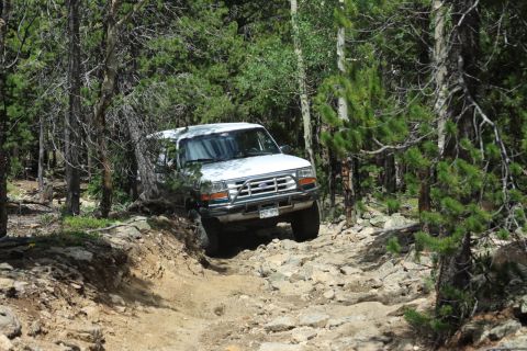 1996 Ford Bronco - White 1996 Ford Bronco driving off-road through a wooded area with rocks.