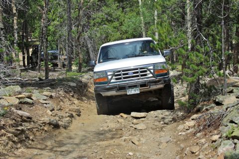 1996 Ford Bronco - A white Ford truck navigates a rough, wooded trail with another vehicle visible in the background.