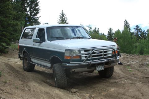 1996 Ford Bronco - A 1996 Ford Bronco with a custom front bumper guard navigates a dirt trail in a wooded area.