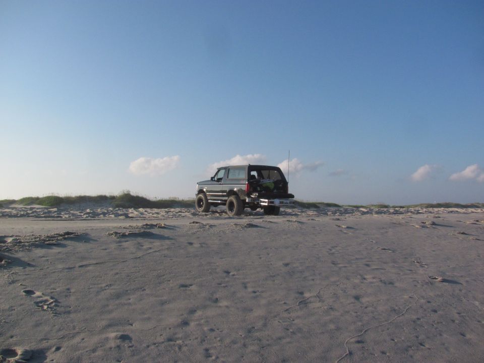 Dark 1995 Ford Bronco parked on a sandy beach with dunes and blue sky