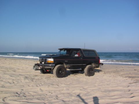 1995 Ford Bronco - A black Ford Bronco sits on the sand near the ocean under a clear blue sky.