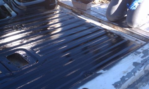 1978 Ford Bronco - A person applies a dark coating to the ribbed floor of a truck bed.