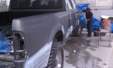 1978 Ford Bronco - A person works on sanding and prepping the body of a gray truck for painting, with blue tarps in the background.