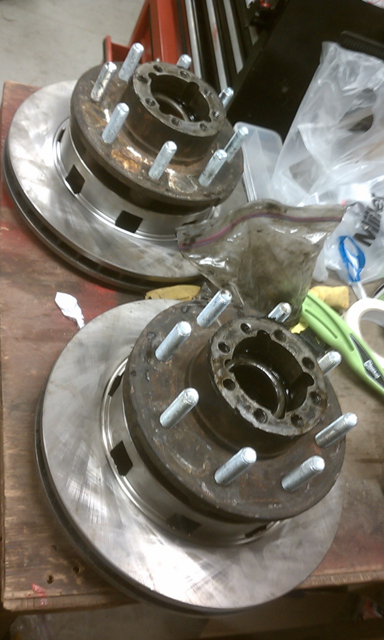 Two rusty brake rotor and hub assemblies with multiple lug studs sit on a wooden surface