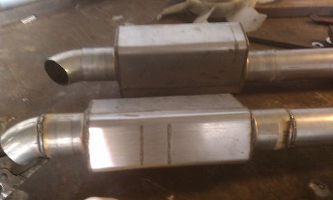 1978 Ford Bronco - Two polished stainless steel oval mufflers with turn-down outlets resting on a metal workbench.