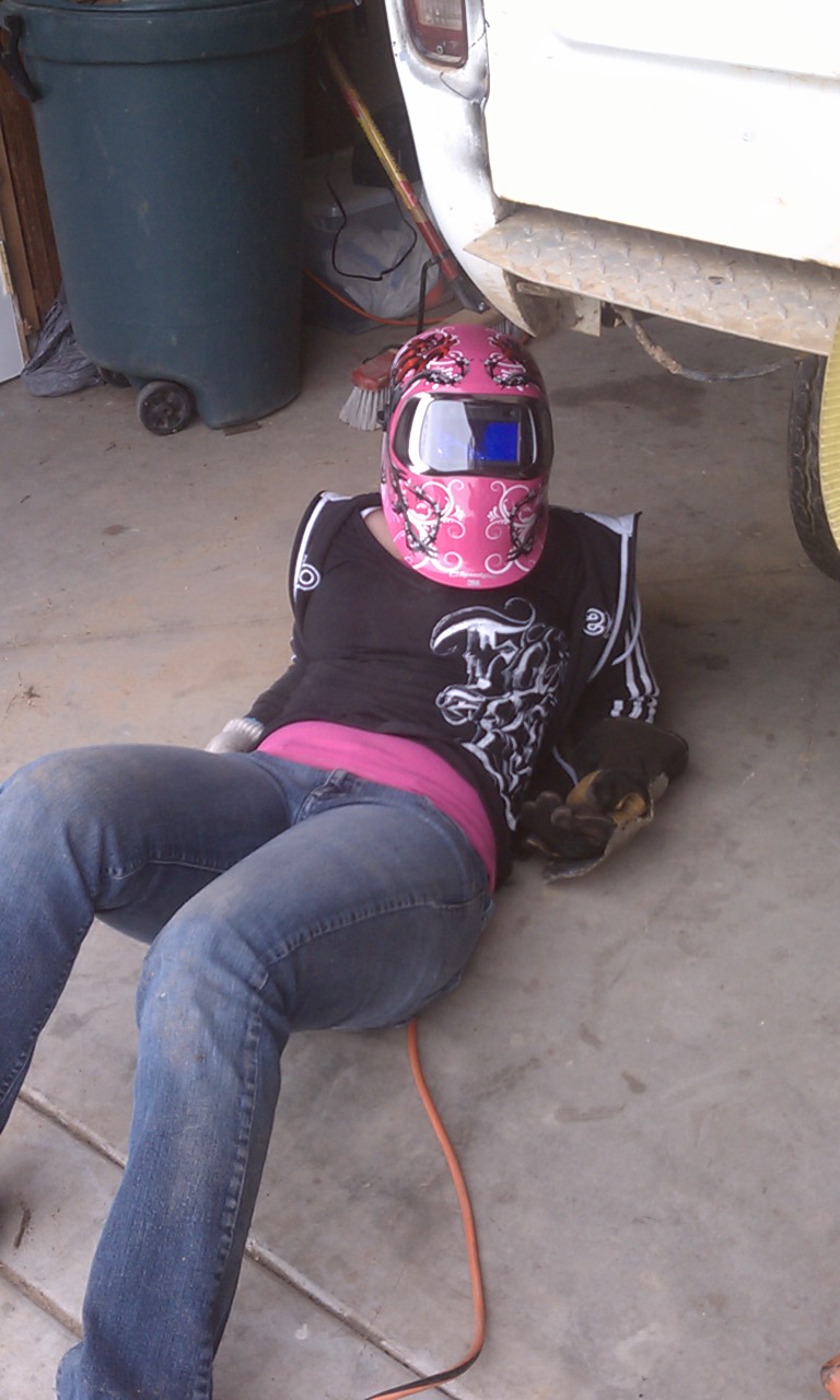 Person in welding mask and jeans sits on concrete floor next to a large trash can and a white vehicle's rear bumper