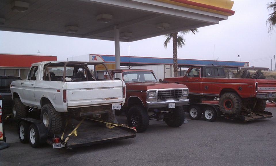 Three lifted trucks parked under a gas station canopy. The truck on the left is a white Ford Bronco on a trailer. A red Ford pickup is on a trailer to the right