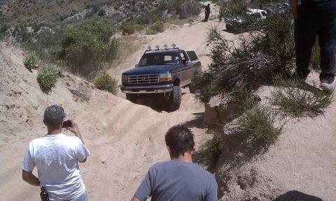 1978 Ford Bronco - A dark blue Ford Bronco navigates a challenging dirt trail, with spectators observing the off-road action.