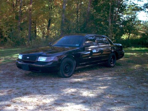 2000 Ford Crown Victoria - A black 2000 Ford Crown Victoria sedan is parked outdoors on a dirt surface with trees behind it.