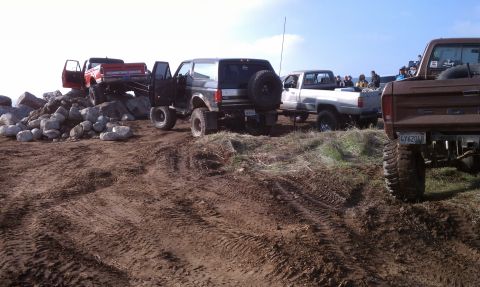 1978 Ford Bronco - Group of lifted 4x4s parked along a rutted dirt ridge at a trail gathering: a black and silver full-size Ford Bronco with a tailgate-mounte…