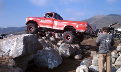 1978 Ford Bronco - Red dentside-era Ford Ranger-trim regular-cab pickup, built as a rock crawler on huge tires and red beadlock wheels, flexing across a bould…