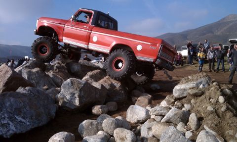 1978 Ford Bronco - Red and white dentside-era Ford Ranger regular-cab pickup climbing nose-up over a rock pile, driver at the wheel, riding on tall mud tires…