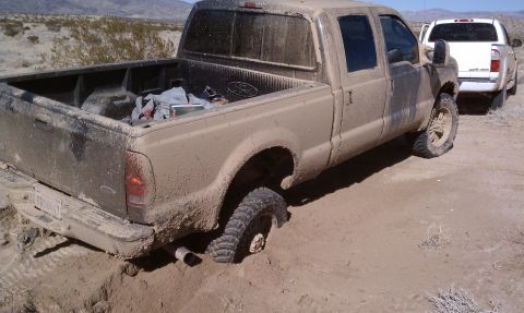 1978 Ford Bronco - Three-quarter rear view of a Ford Super Duty crew cab pickup coated head-to-toe in dried tan mud, sitting in soft desert sand with mud-terr…