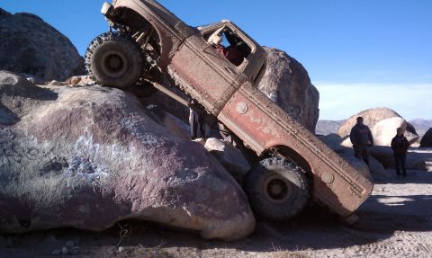 1978 Ford Bronco - A mud-caked, weathered full-size Bronco-bodied rock crawler climbs a huge boulder at a steep nose-up angle in a desert rock garden, front w…