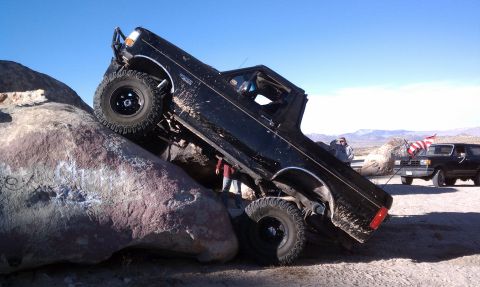 1978 Ford Bronco - Black lifted Ford pickup climbing steeply nose-up over a large granite boulder in the desert, front tires airborne on the rock face with th…