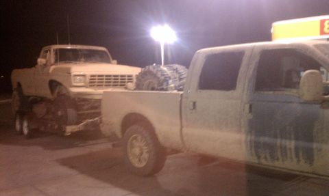 1978 Ford Bronco - Night shot at a lit fuel stop of a dust-covered crew-cab Super Duty pickup towing a trailer loaded with a stripped, mud-caked older Ford tr…
