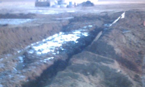 1978 Ford Bronco - Blurry night shot of a water-filled mud pit dug into a dirt course, with a rutted berm and a marker stake alongside it; trucks and spectato…