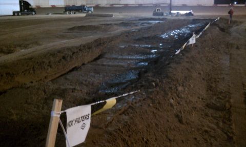 1978 Ford Bronco - A freshly cut mud bog lane at an indoor/covered event venue at night, the water-filled trench lined with WIX Filters banner flags on wooden…