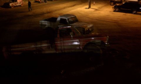 1978 Ford Bronco - Dark, blurry nighttime shot of a red lifted pickup on oversized mud tires parked on a dirt lot, with a mud-splattered white full-size Bronc…