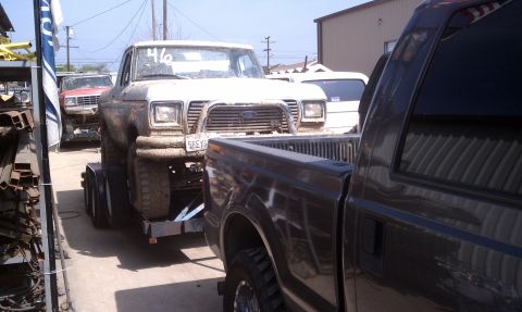 1978 Ford Bronco - Front view of a white square-body Ford pickup, mud-splattered along the fenders and stripped of doors and hood trim, loaded on a tandem-axl…