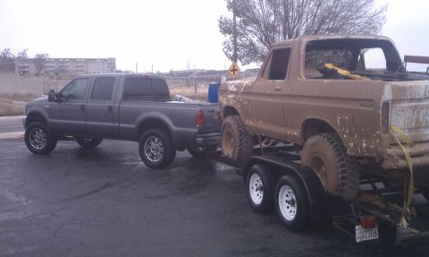 1978 Ford Bronco - A mud-caked square-body Ford pickup with no doors, glass or tailgate strapped down with yellow ratchet straps on a tandem-axle flatbed trai…