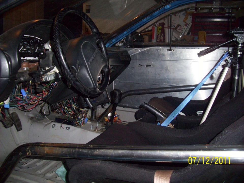 Interior of a car with racing seats, roll cage, and a mess of wires under the dashboard. Steering wheel has 'PROBE' on it
